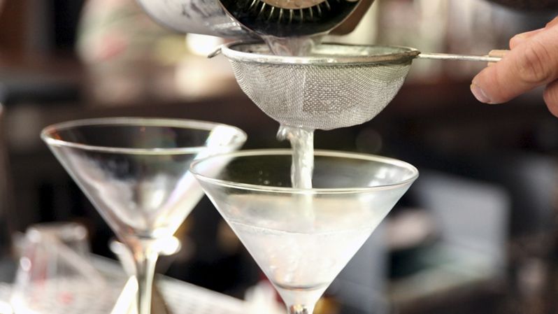 a bartender pouring martinis into chilled glasses through a small wire mesh strainer.